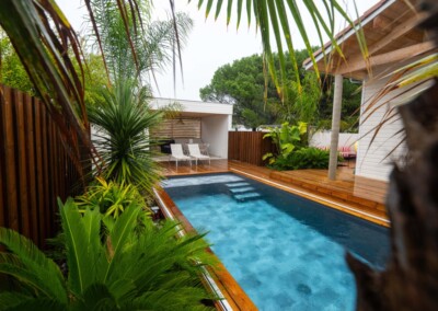 Piscine rectangulaire entourée de végétation tropicale, avec terrasse en bois et chaises blanches sous un abri ouvert.