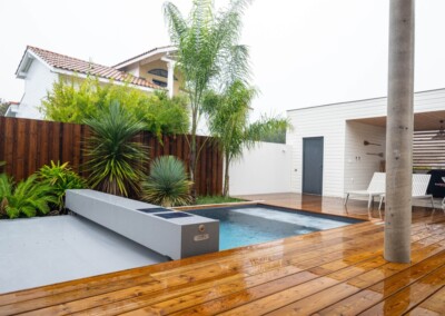 Piscine moderne entourée de plantes tropicales, terrasse en bois mouillé, maison avec toit en tuiles et pool house blanc.