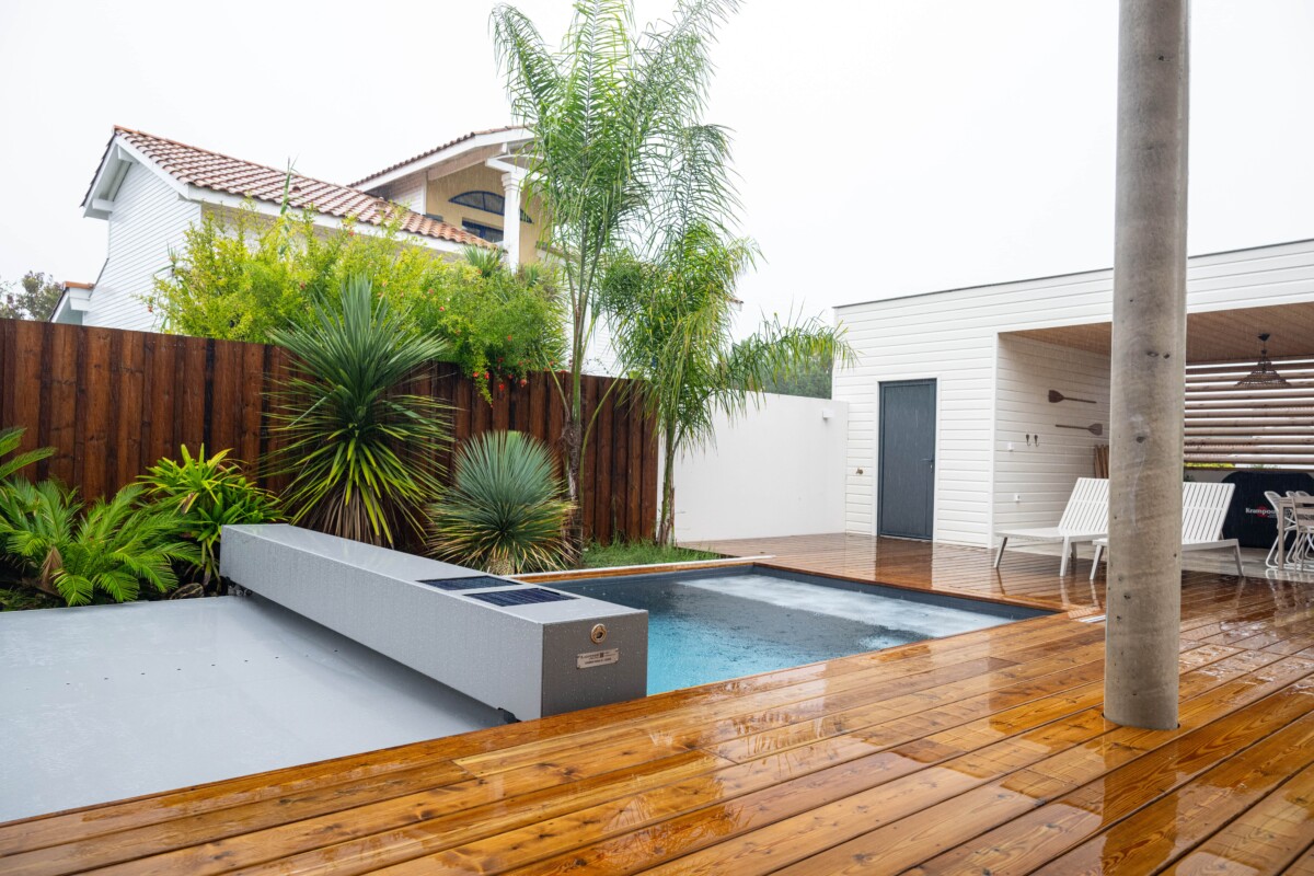 Une piscine entourée de palmiers et de plantes, avec une terrasse en bois mouillée et un bâtiment blanc en arrière-plan.