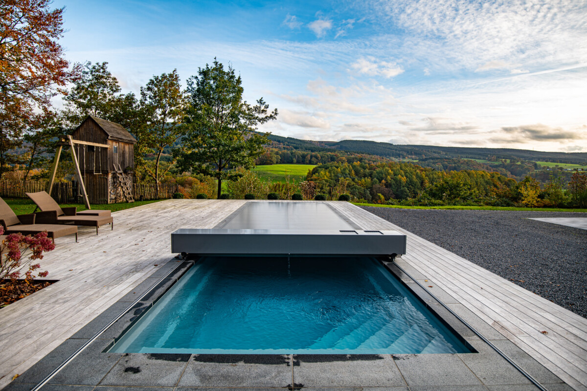Piscine couverte par une structure grise sur une terrasse en bois, avec des arbres et des collines en arrière-plan sous un ciel partiellement nuageux.