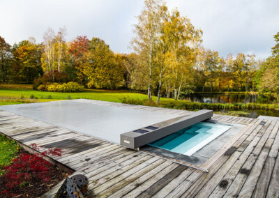 Piscine rectangulaire équipée d’une couverture Coverseal grise, entourée d’une terrasse en bois, avec un grand jardin verdoyant et un étang en arrière-plan.