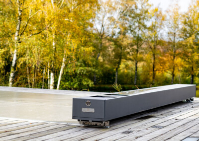 Vue rapprochée du mécanisme Coverseal posé sur une terrasse en bois, protégeant une piscine, avec des arbres aux couleurs automnales en arrière-plan.