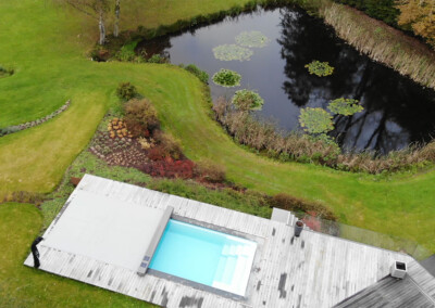 Vue aérienne d’une piscine avec couverture Coverseal et terrasse en bois, installée dans un jardin paysager à côté d’un grand étang.