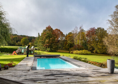 Piscine turquoise entourée d’une terrasse en bois, avec aire de jeux pour enfants et jardin aux couleurs d’automne en arrière-plan.